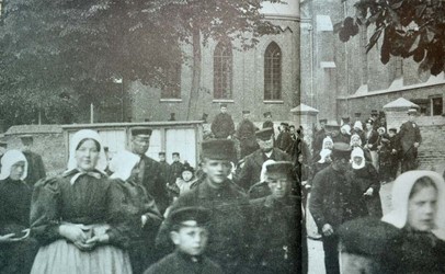 <p>Het uitgaan van de kerk in 1902, gefotografeerd vanuit het zuidoosten. Tussen de kerkgangers door is de ommuring te zien, die aan deze zijde in 1908 werd vervangen door een ijzeren hek. </p>
